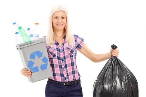 A young woman with long blonde hair, dressed in a short-sleeved pink and blue checkered shirt and dark trousers, stands outdoors against a plain white background. She is smiling and holding a small grey recycling bin filled with clear plastic bottles and containers, with a blue recycling symbol on the front. In her other hand, she carries a large black plastic refuse bag. The scene appears staged, with no specific environmental surroundings visible, emphasizing her role in rubbish collection or disposal, relevant to services offered by Office Clearance Chigwell in the local area. The bright lighting and clear focus highlight the materials and the action of waste handling, aligning with professional rubbish removal services in the Chigwell area.