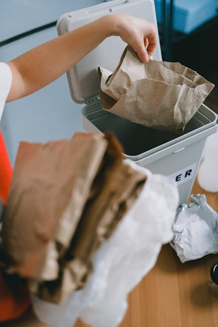The image shows a person emptying a brown paper bag into a white rubbish bin with a black liner, placed on a light-colored surface indoors. The individual’s arm is visible, wearing a white short-sle