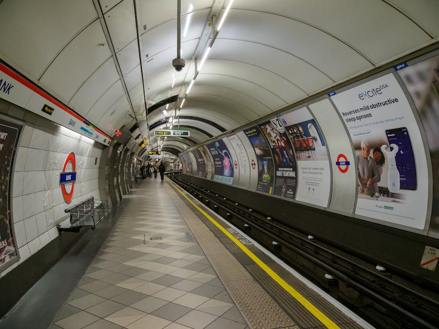 A London Underground train with a white body and orange and blue accents is stationary at an elevated train station platform, which features a grey tiled surface with yellow tactile paving along the e