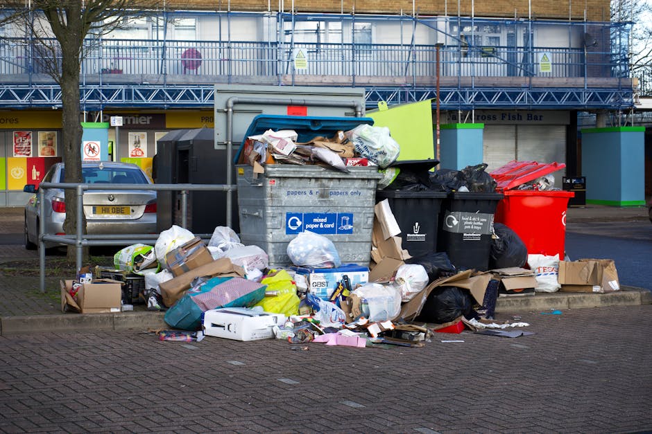 A roadside collection point showing a large pile of mixed household waste and recyclable materials situated in a parking area adjacent to a commercial building. The pile includes various black plastic