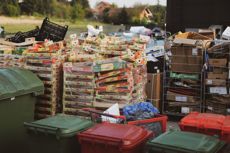 A roadside collection point showing a large pile of mixed household waste and recyclable materials situated in a parking area adjacent to a commercial building. The pile includes various black plastic