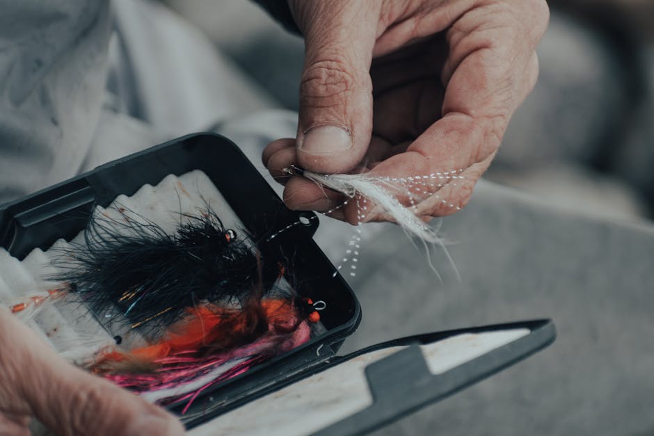 A person’s hand is seen holding a small, clear fishing hook with a white feather attached, positioned above an open, rectangular tackle box containing a collection of feathers, synthetic fibers, and o