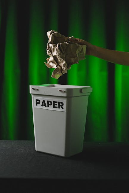 A large collection of mixed household waste and paper and cardboard waste is piled up on a paved sidewalk area, adjacent to a parking space. The waste includes various black and green plastic rubbish
